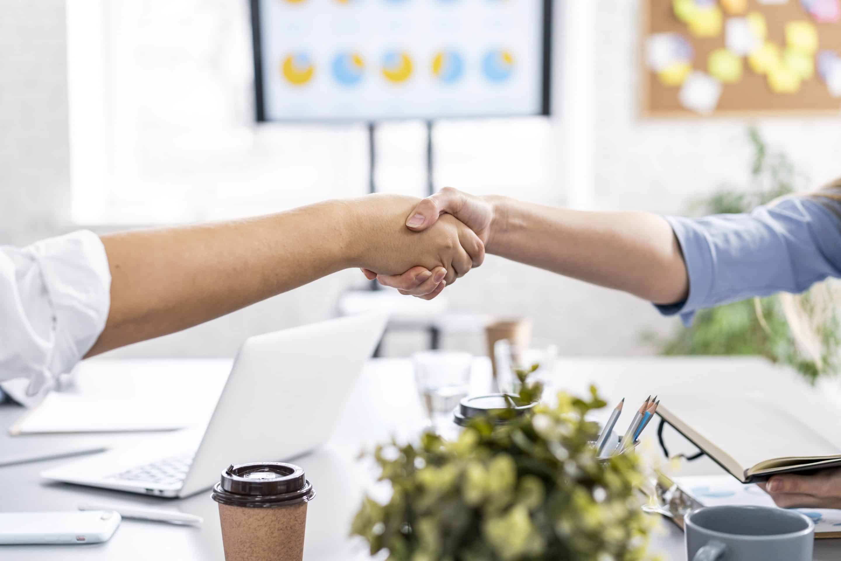 Deux personnes se serrent la main au-dessus d’un bureau dans un environnement de travail moderne, avec un ordinateur portable, des documents et des tasses en arrière-plan.